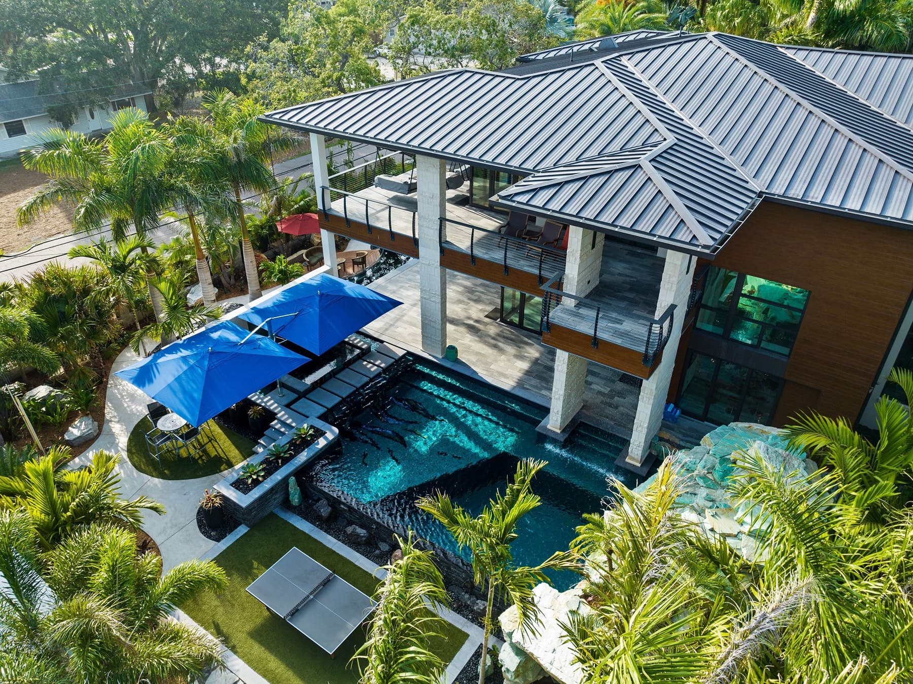 Drone view over metal-roof residence, blue umbrellas and grotto pool below
