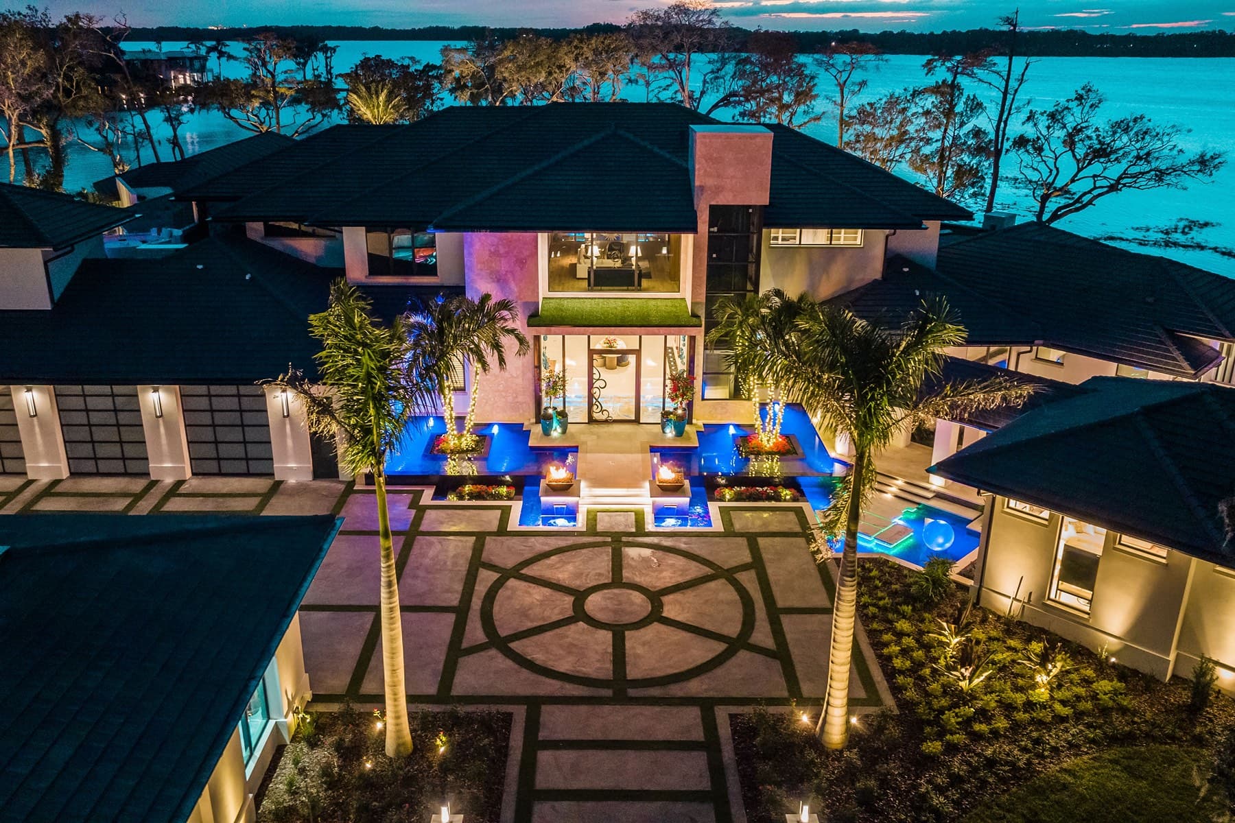 Twilight aerial of grand estate courtyard with palms and lake beyond