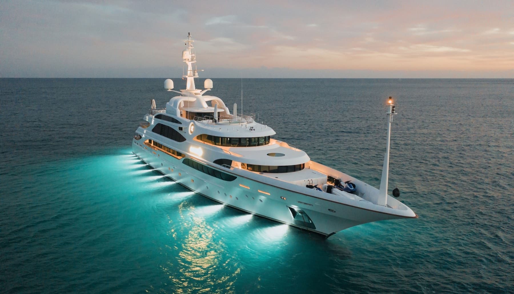Superyacht glowing at dusk with turquoise underwater lights on dark sea