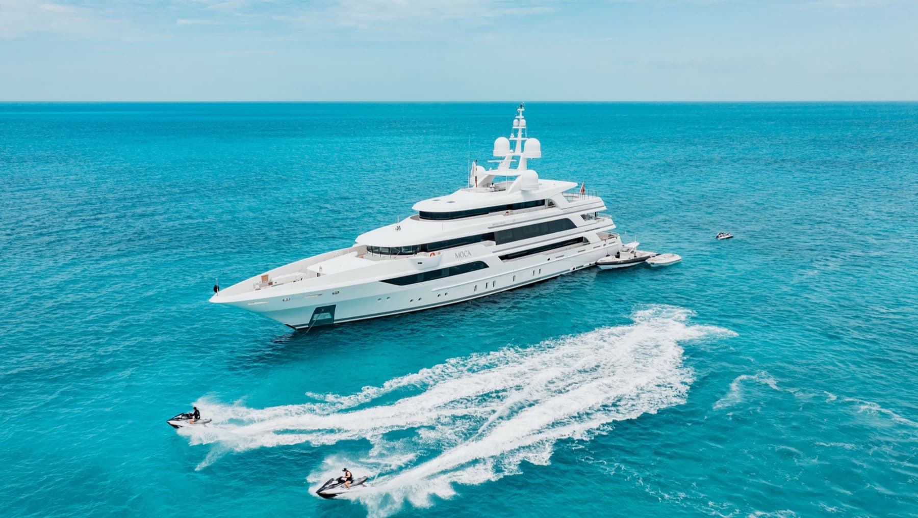 Aerial of white superyacht at anchor with jet skis carving turquoise water