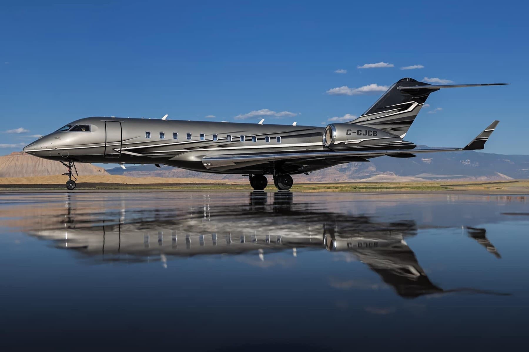 Gunmetal Bombardier Global jet mirrored in wet tarmac against desert mountains