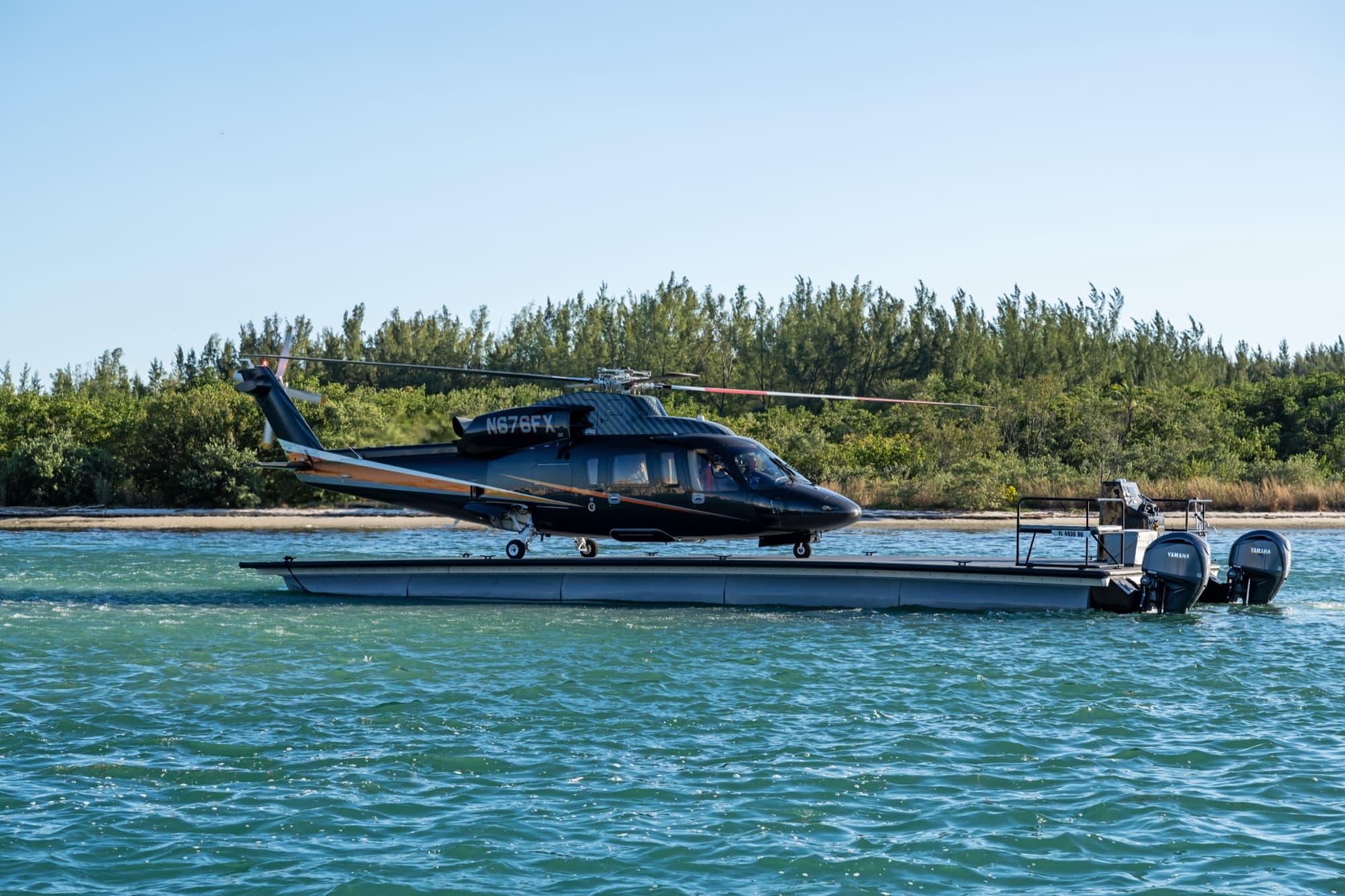 Black Sikorsky S-76 on a waterfront helipad with a yacht beyond